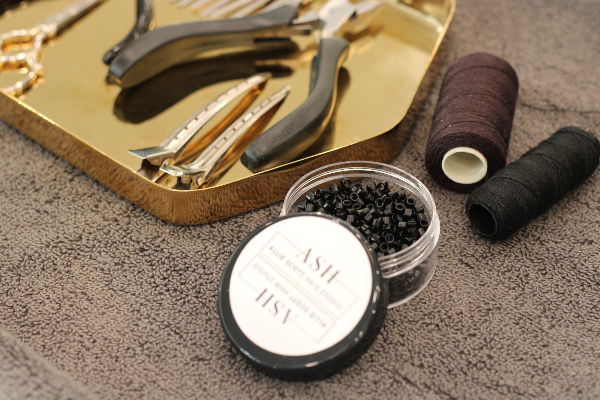 Hair extension tools and materials including pliers, wire, and a container of beads on a textured surface.